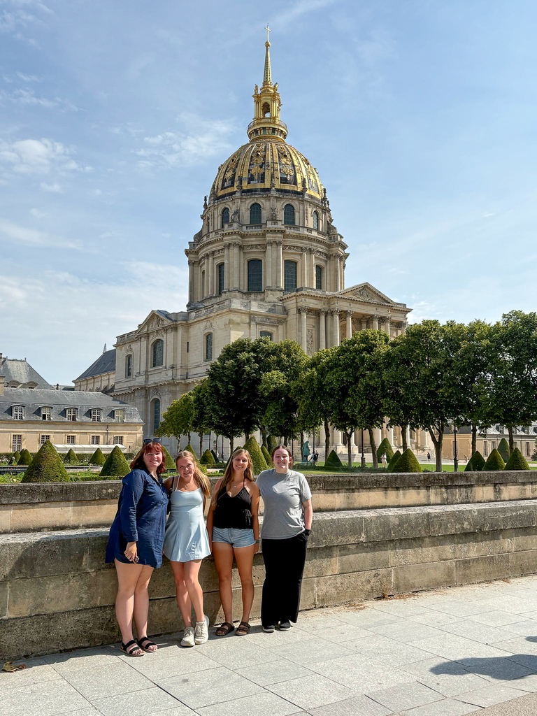 Four people standing and smiling in front of the golden dome of Les Invalides in Paris on a sunny day, with manicured gardens and historic architecture in the background.