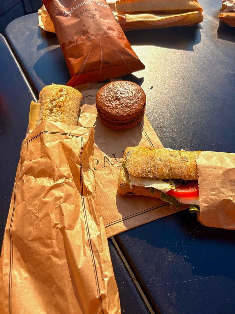 An outdoor café table with fresh French sandwiches on baguettes, potato chips, and a chocolate macaron resting on brown paper wrappers in warm afternoon sunlight.