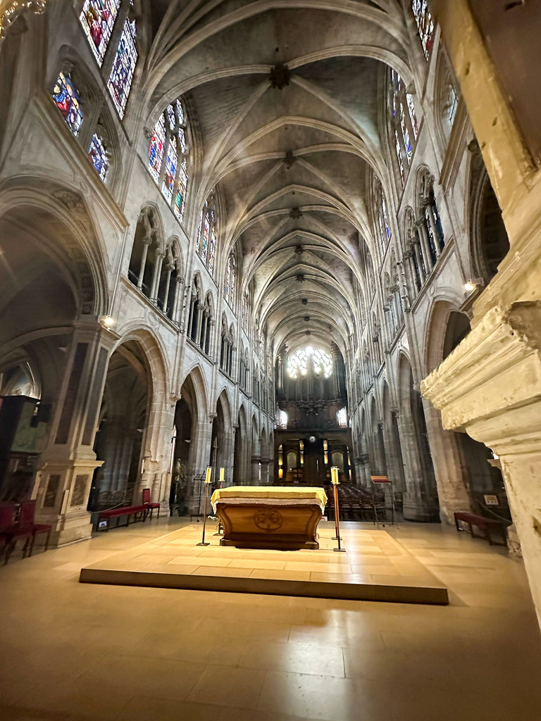 Interior of a grand Gothic cathedral with soaring vaulted ceilings, tall stone columns, stained glass windows, and a central altar illuminated by natural light streaming through a large rose window.