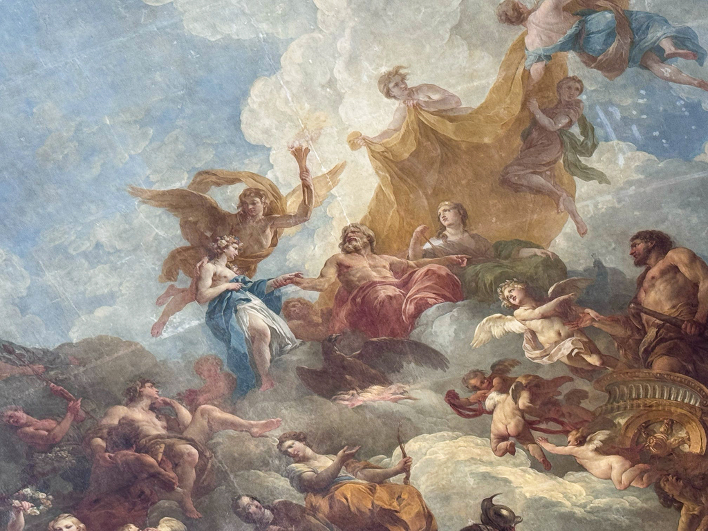A Baroque ceiling fresco depicting mythological figures, angels, and clouds painted in soft blues, golds, and earth tones inside an ornate palace room.