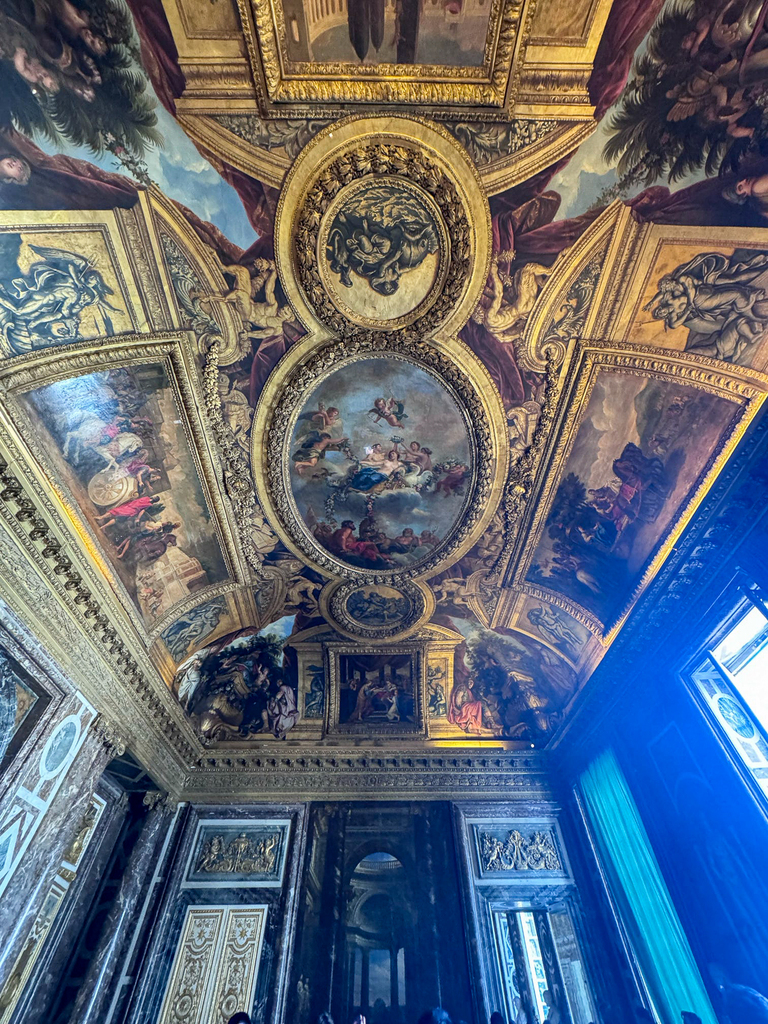 An elaborately decorated palace ceiling featuring gilded moldings, framed paintings, and ornate architectural details illuminated by natural light from tall windows.