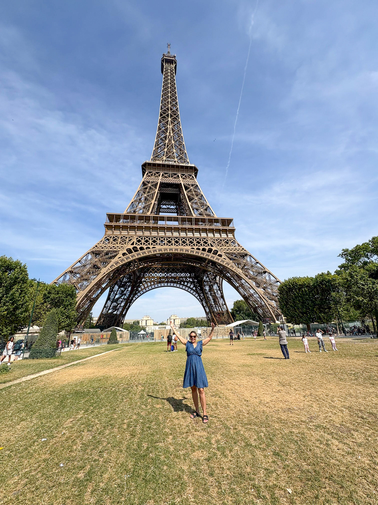 The Eiffel Tower viewed from the Champ de Mars, framed against a bright blue sky as visitors walk across the lawn and pathways beneath the landmark.