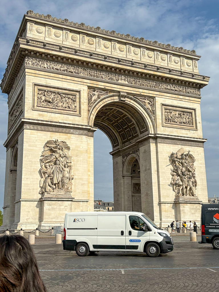 The Arc de Triomphe in Paris viewed from the front, showcasing detailed stone carvings and sculptures. A white delivery van and pedestrians are visible in the foreground.