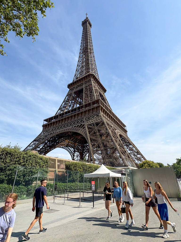 The Eiffel Tower photographed from below on a clear blue day, highlighting its intricate iron lattice structure with visitors walking nearby.