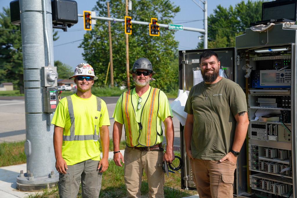 Three road crew members stand beside an open traffic-signal control cabinet at the new intersection of State Route 131 and Eagles Watch Way.