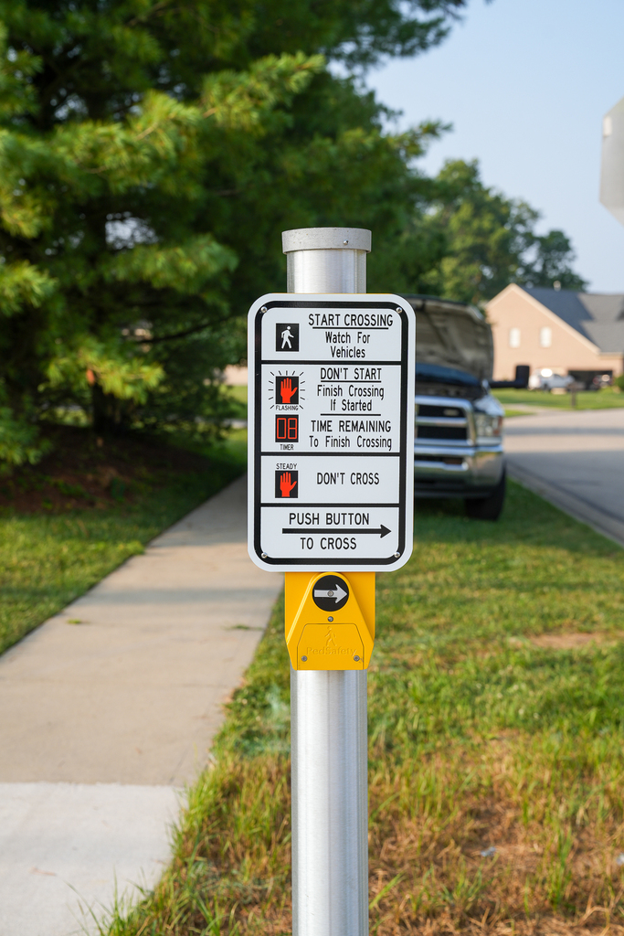 A pedestrian crossing station features a push button and sign explaining the walk signal, flashing countdown and “Don’t Cross” signal.