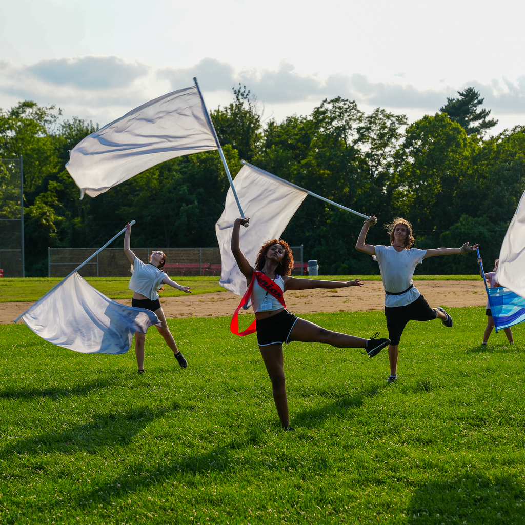 Color guard students perform with large white flags while holding balanced poses on a grassy field.