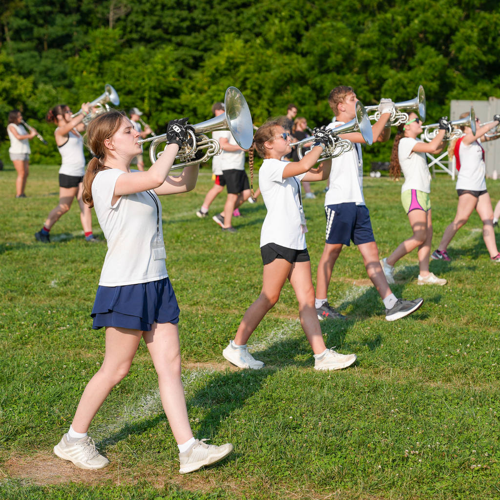 Marching band students play silver brass instruments while stepping forward together during an outdoor rehearsal.