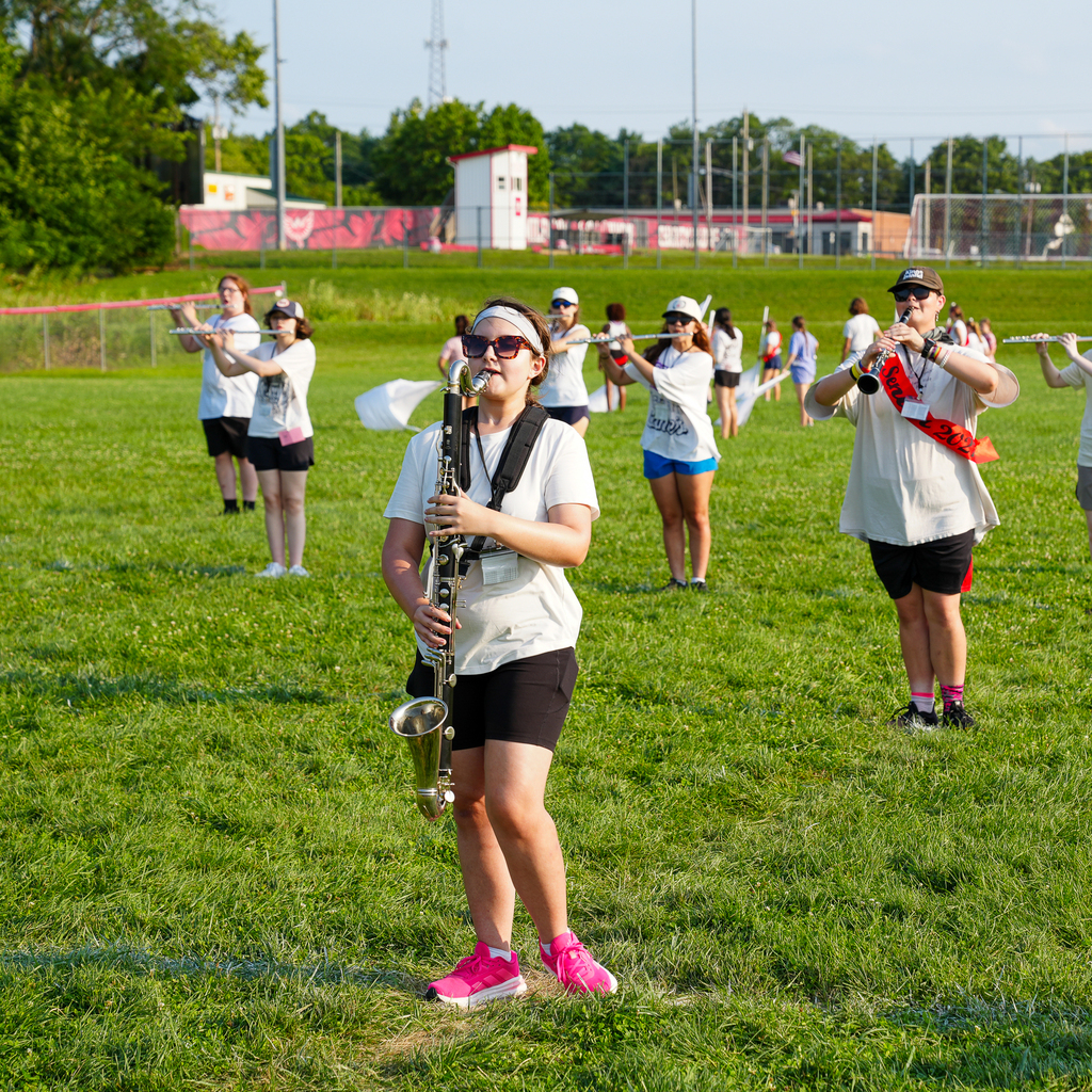 A student plays a large clarinet in the foreground while woodwind musicians and color guard members rehearse behind her.