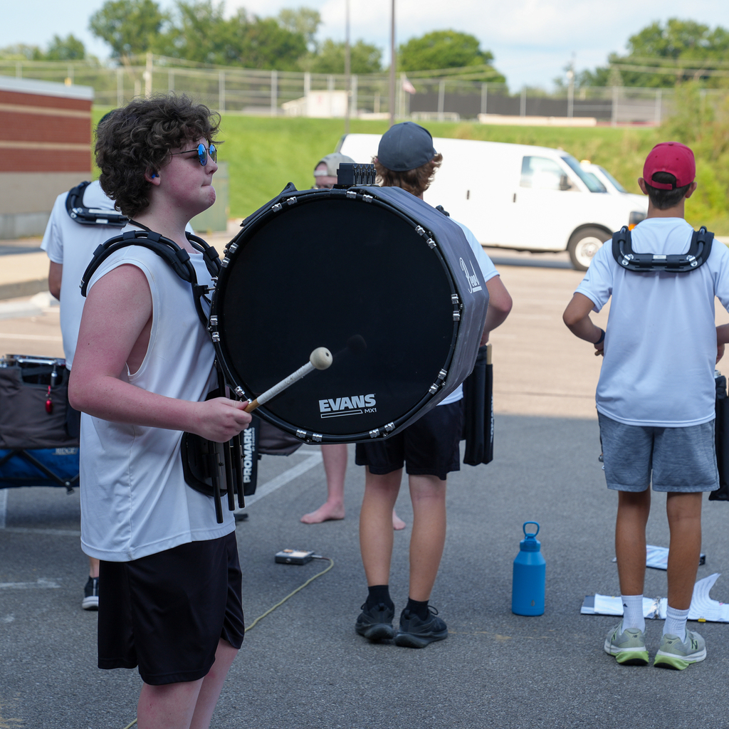 A Milford marching band student carries a large bass drum during an outdoor percussion rehearsal while other members stand nearby.