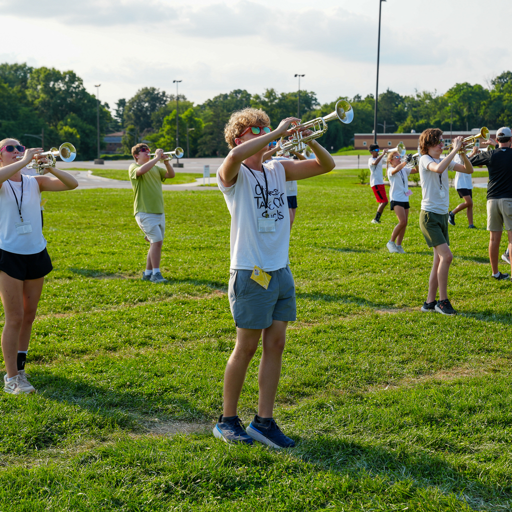 Marching band students practice trumpet music and drill positions in formation on a grassy field.