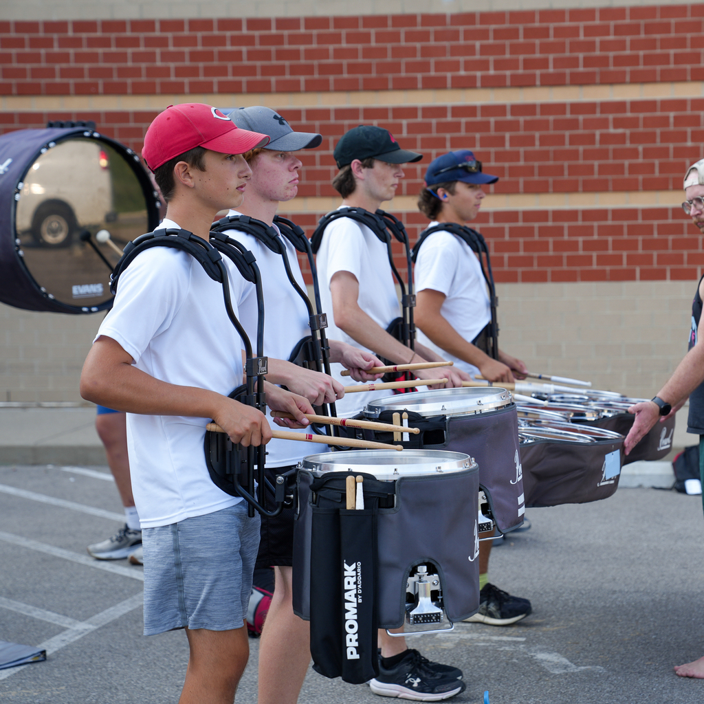 Four marching band students stand in a row with snare drums as they practice together outside the school.