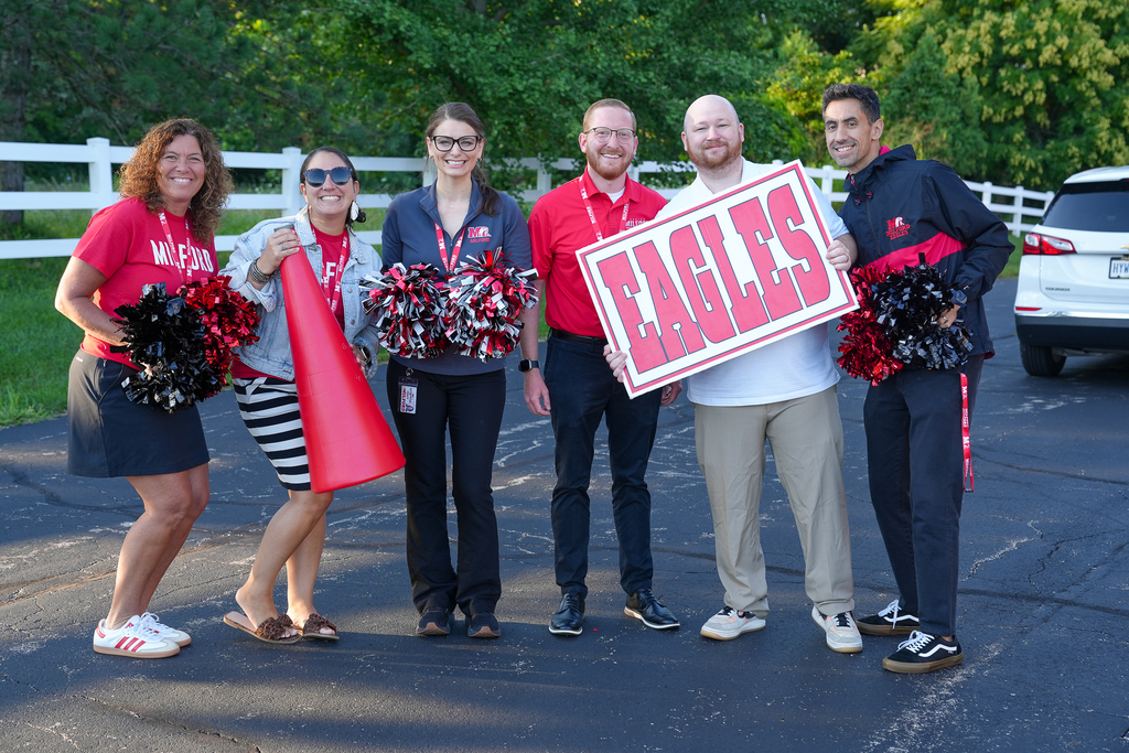 Six Milford staff members pose outdoors with red, black and white pom-poms, a red megaphone and a large “Eagles” sign.