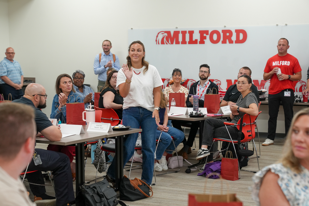 A Milford University participant stands and waves as colleagues seated around classroom tables applaud. A large Milford logo is displayed behind the group.