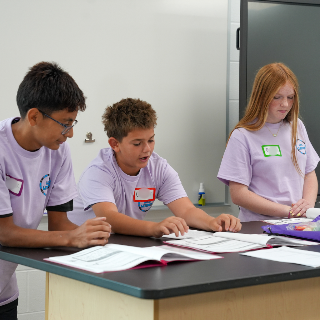 Three Milford Middle School WEB leaders sit at the front of a classroom leading an activity for incoming sixth-grade students. The student leaders reference activity materials while helping guide discussions and answer questions.