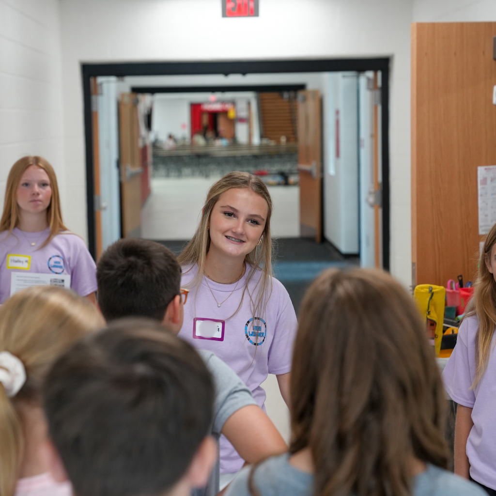 Students gather in a hallway at Milford Middle School during WEB Orientation as a WEB leader in a light purple T-shirt speaks to a small group of incoming sixth graders. Other WEB leaders stand nearby, helping welcome students as they begin exploring the building.