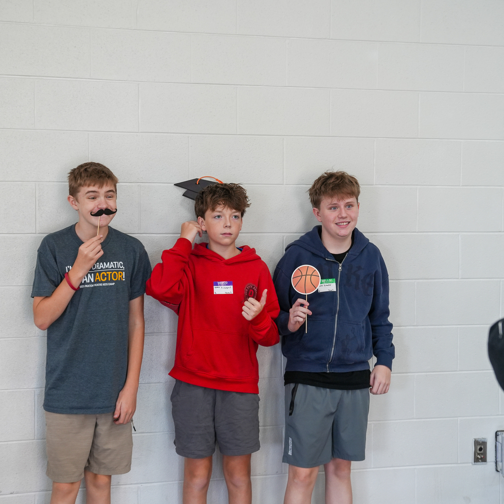Three Milford High School freshmen pose with fun photo booth props, including a mustache, graduation cap, and basketball, during Freshman Orientation.