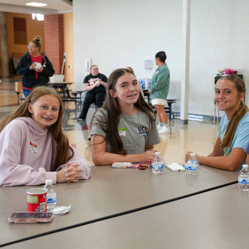 Three Milford High School freshmen sit together at a cafeteria table during Freshman Orientation, smiling at the camera with water bottles and snacks in front of them as they get to know one another.