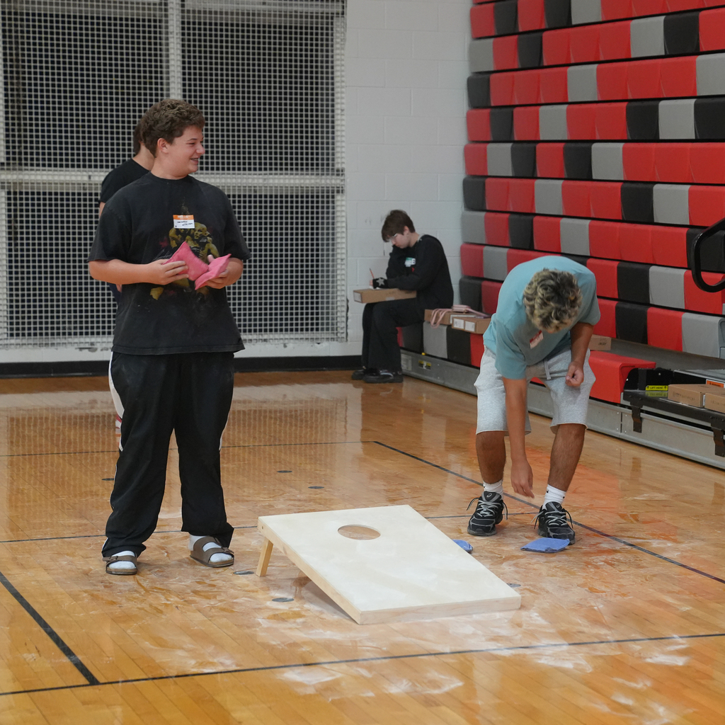Two Milford High School freshmen play cornhole in the gym during Freshman Orientation while other students participate in activities in the background.