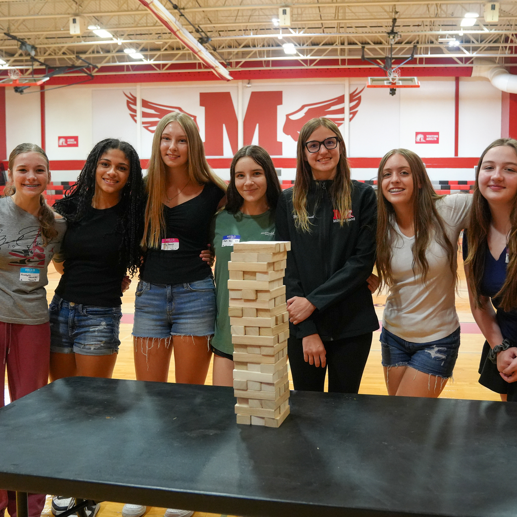 Seven Milford High School students stand together behind a giant Jenga tower in the gymnasium during Freshman Orientation. The group smiles for the camera with the large Milford "M" displayed on the wall behind them.