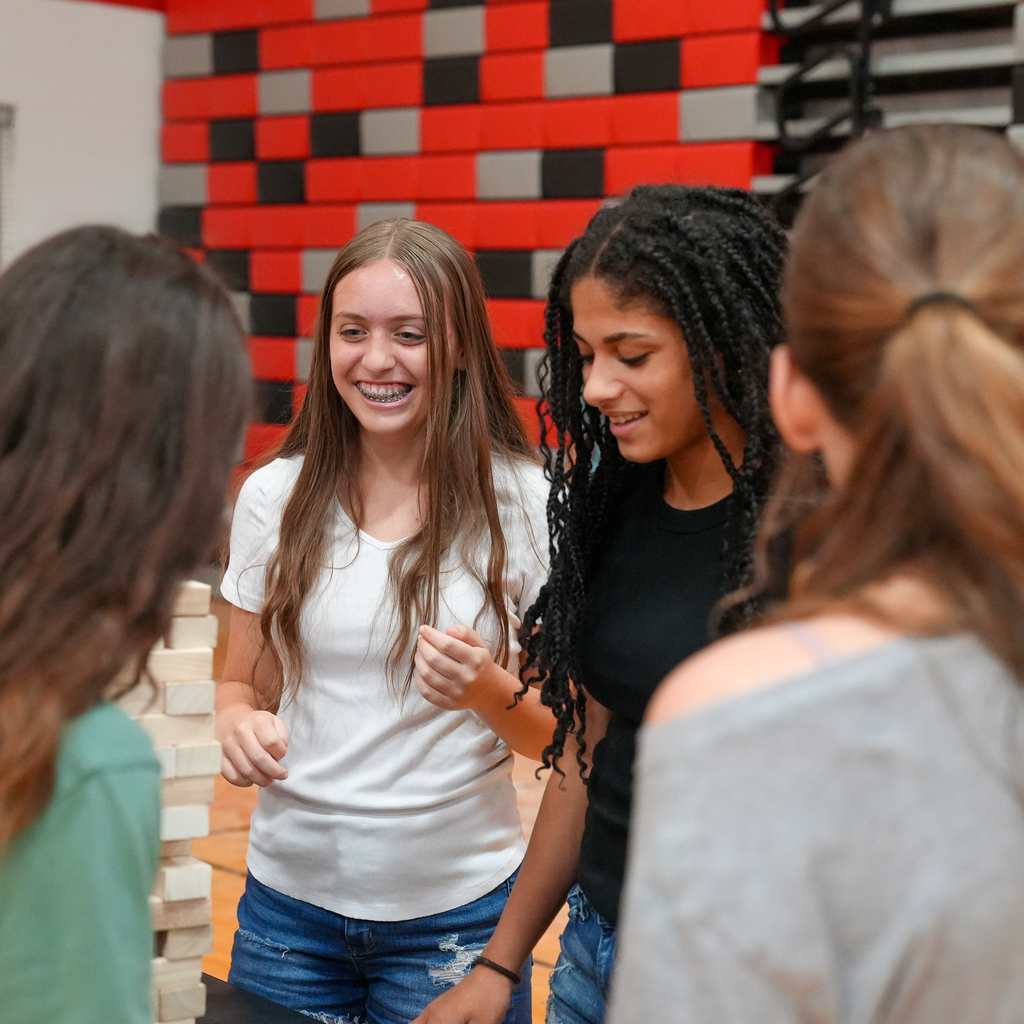 A group of Milford High School freshmen gathers around a giant Jenga game during Freshman Orientation. Two students smile as they prepare for the next move while classmates watch and cheer them on.