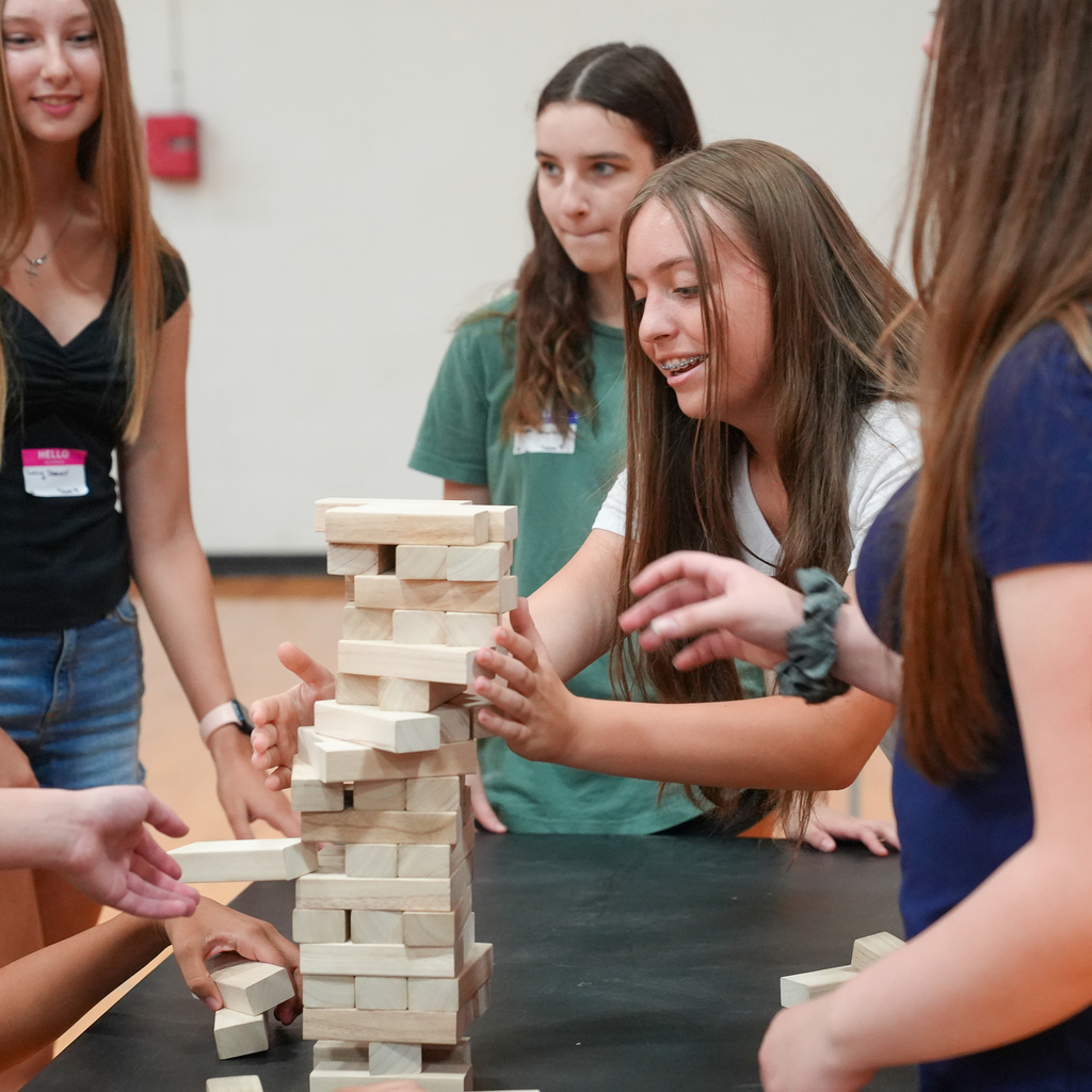 A Milford High School freshman carefully removes a wooden block from a giant Jenga tower while classmates gather around the table, watching and encouraging her during an orientation activity.
