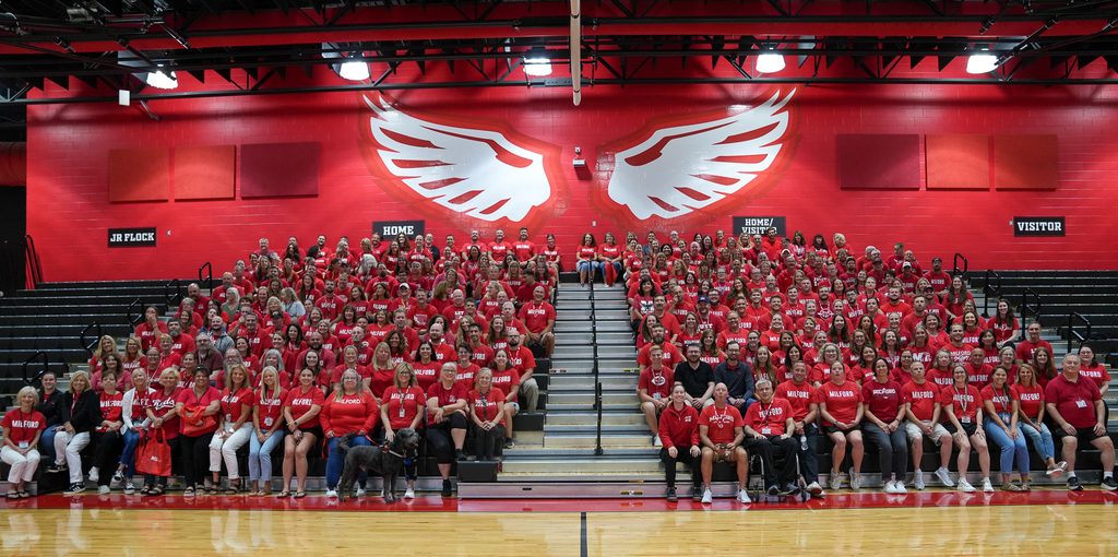 A large group of Milford staff members, most wearing red, poses on the high school gymnasium bleachers beneath a mural of white eagle wings.