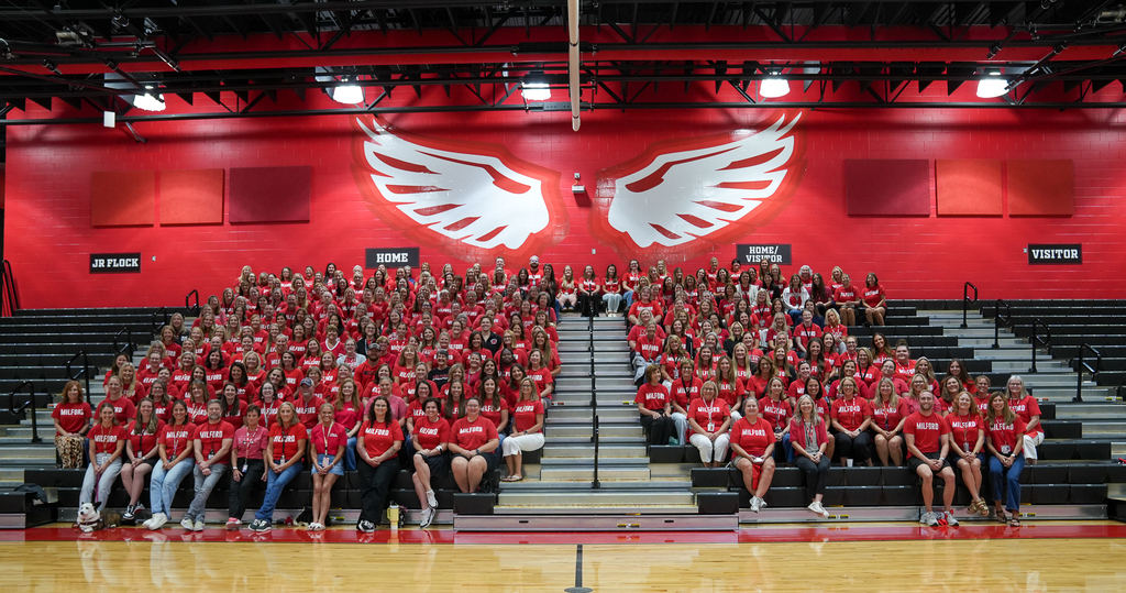 Hundreds of Milford staff members dressed in red smile for a group photo on the high school gymnasium bleachers beneath a mural of white eagle wings.