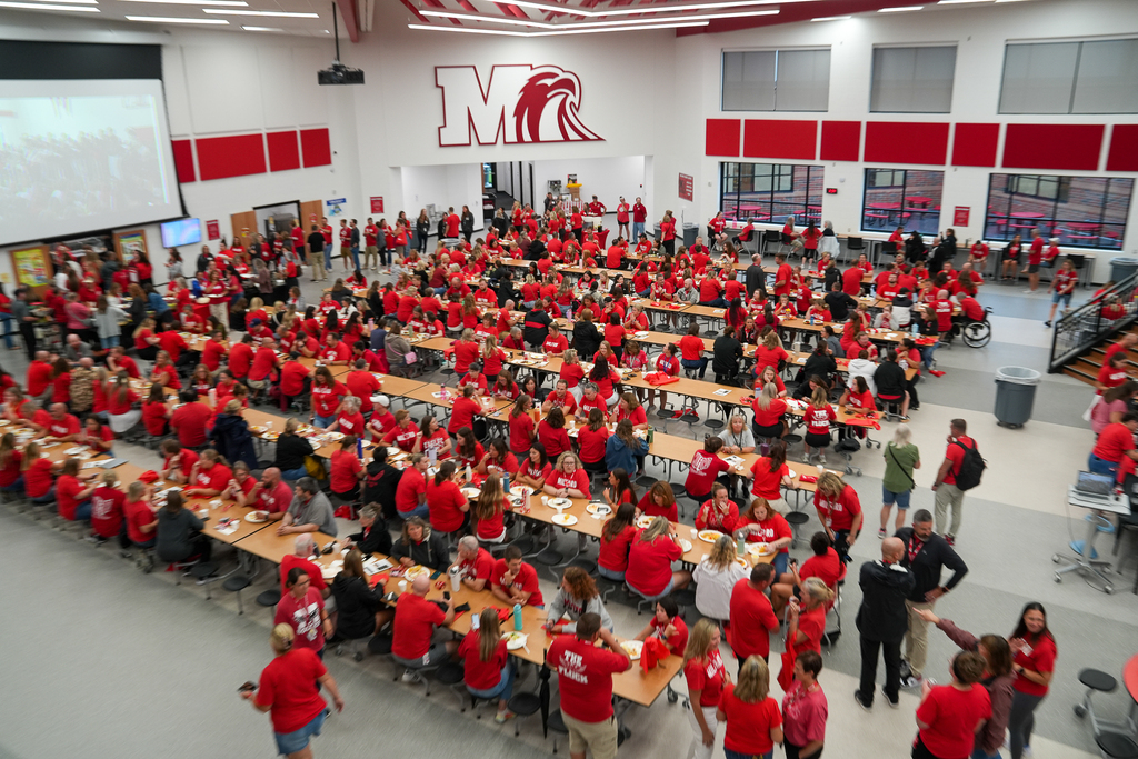 An overhead view shows hundreds of Milford staff members, many wearing red, gathered around cafeteria tables for the annual staff convocation.
