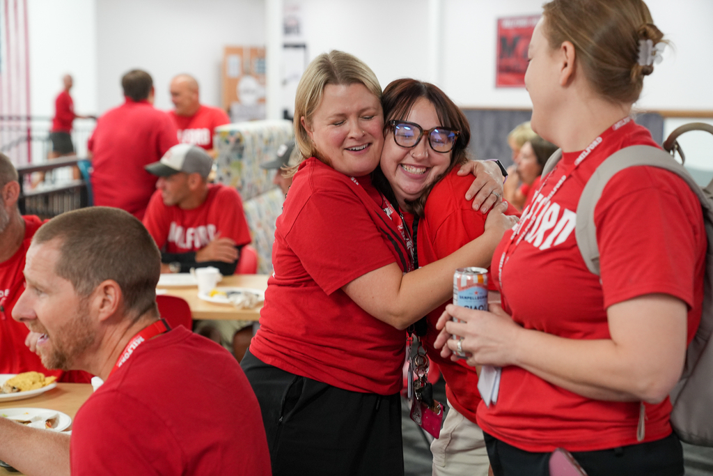 Two Milford staff members smile and hug during convocation as other colleagues gather nearby.