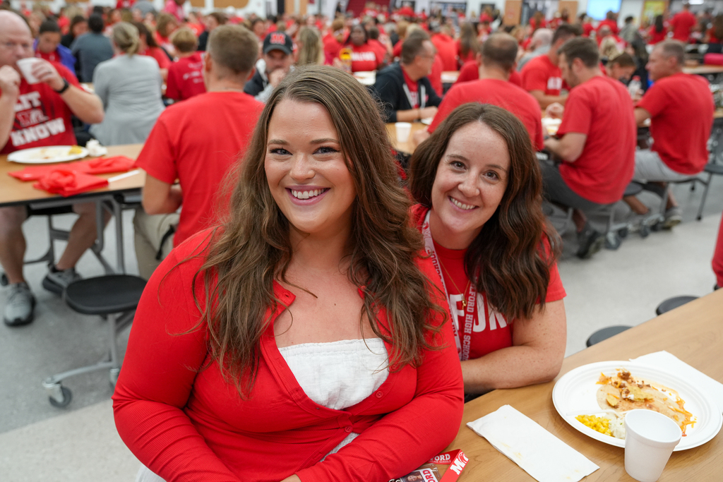 Two Milford staff members smile for a photo while seated in the crowded high school cafeteria during convocation.