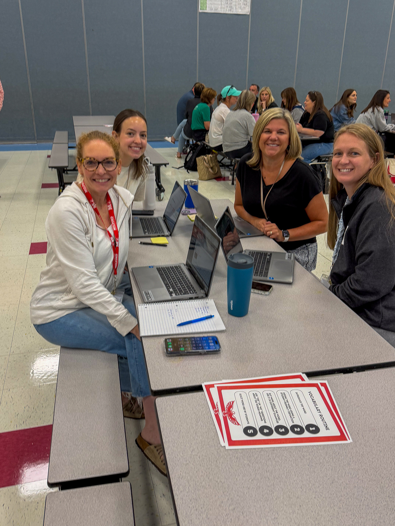 Four smiling staff members sit together at a cafeteria table with laptops and professional development materials.
