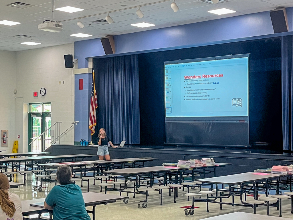 A staff member presents information about Wonders instructional resources to colleagues gathered in the school cafeteria.