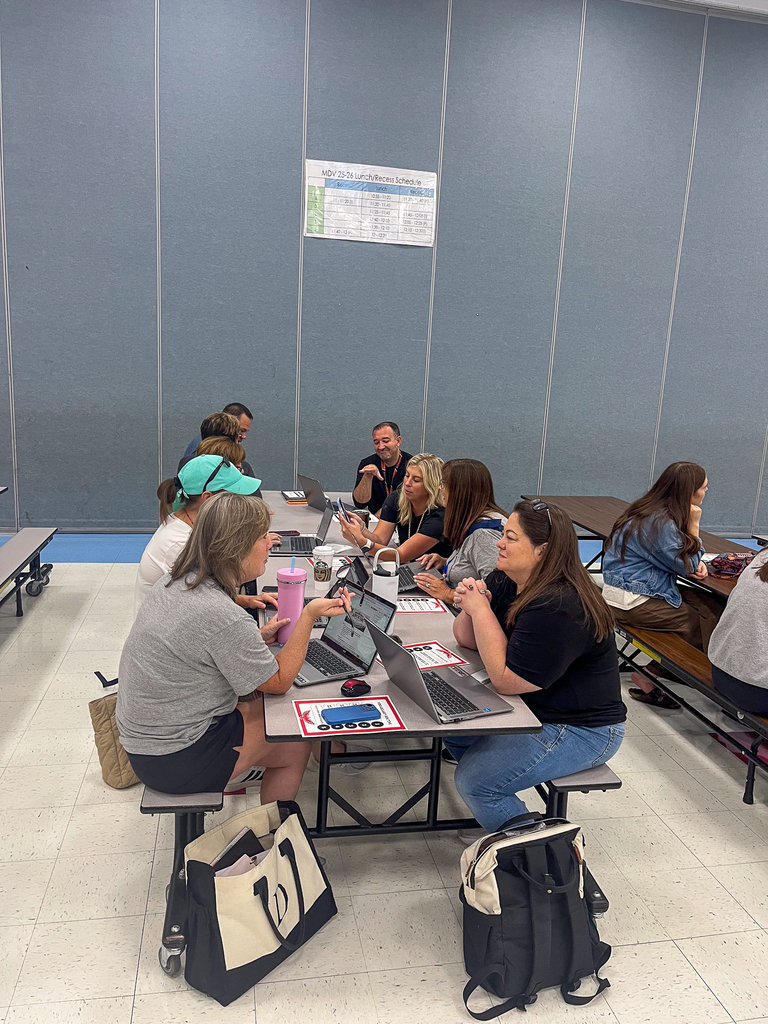 Staff members collaborate around cafeteria tables while working on laptops and discussing instructional materials.