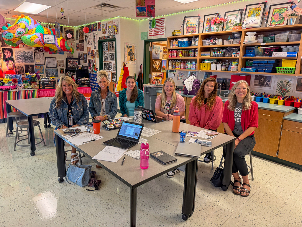 Six smiling staff members sit around tables with laptops, notebooks and classroom materials in a colorful art room.