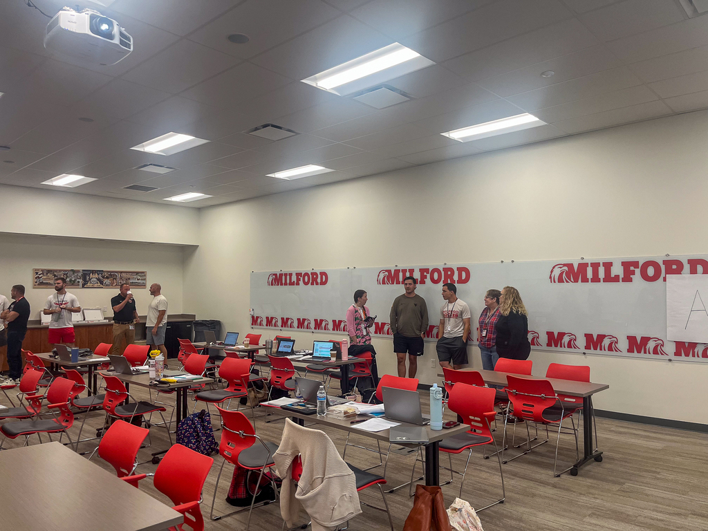 Milford staff members gather in a classroom for professional development, standing in groups among tables, laptops and red chairs.