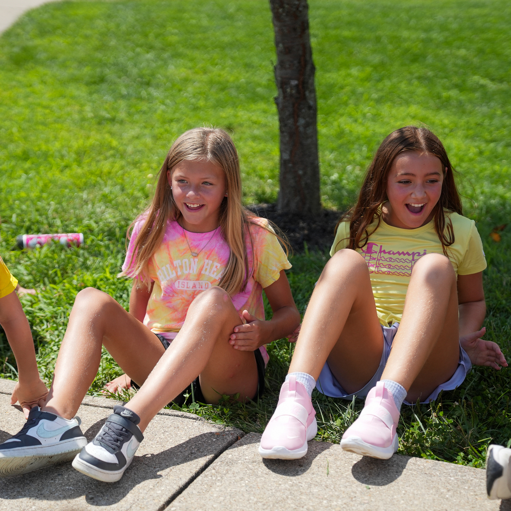 Two McCormick Elementary students smile and laugh while seated on the grass during an outdoor PBIS celebration.