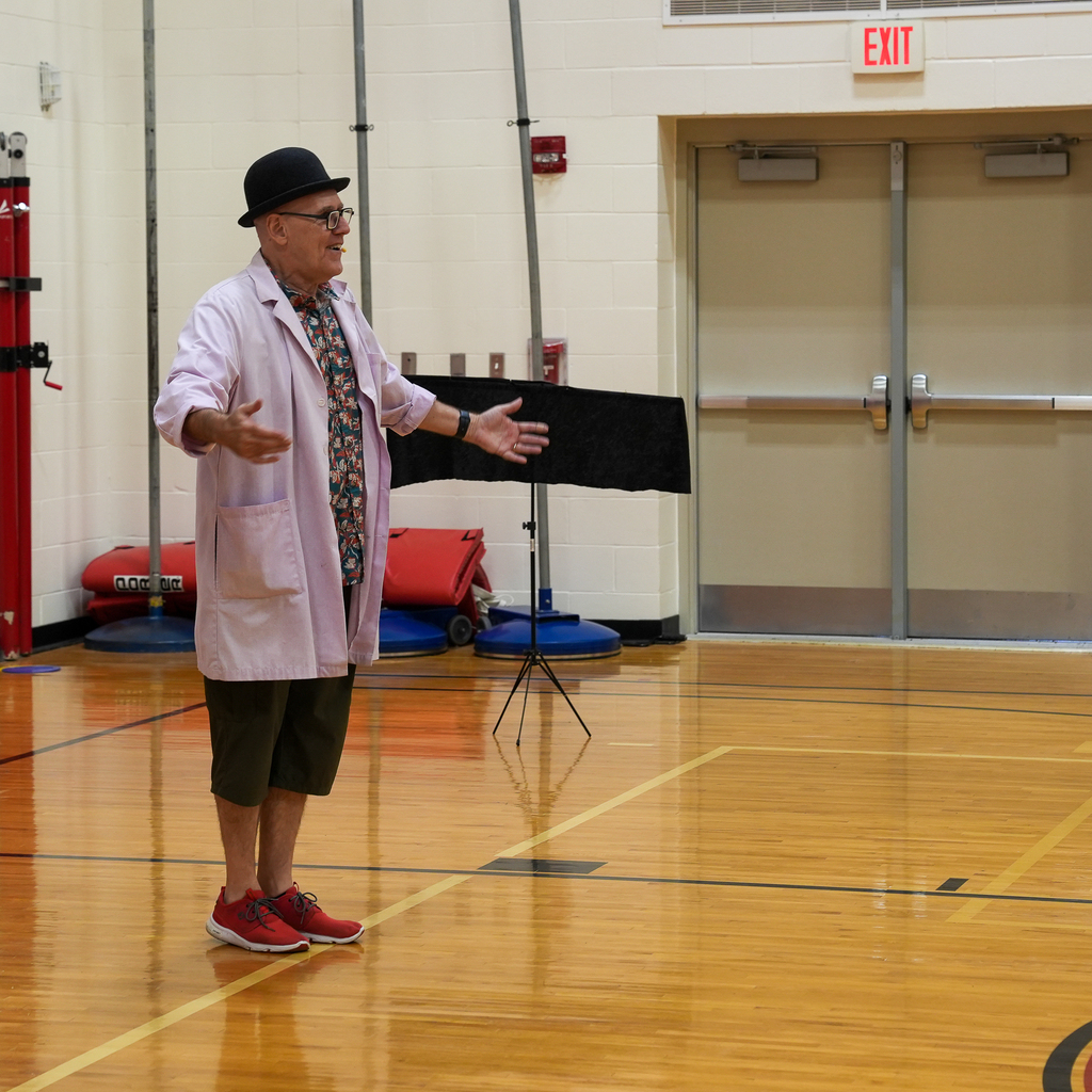 A performer wearing a bowler hat and lab coat speaks to students during an assembly in the Boyd E. Smith gymnasium.