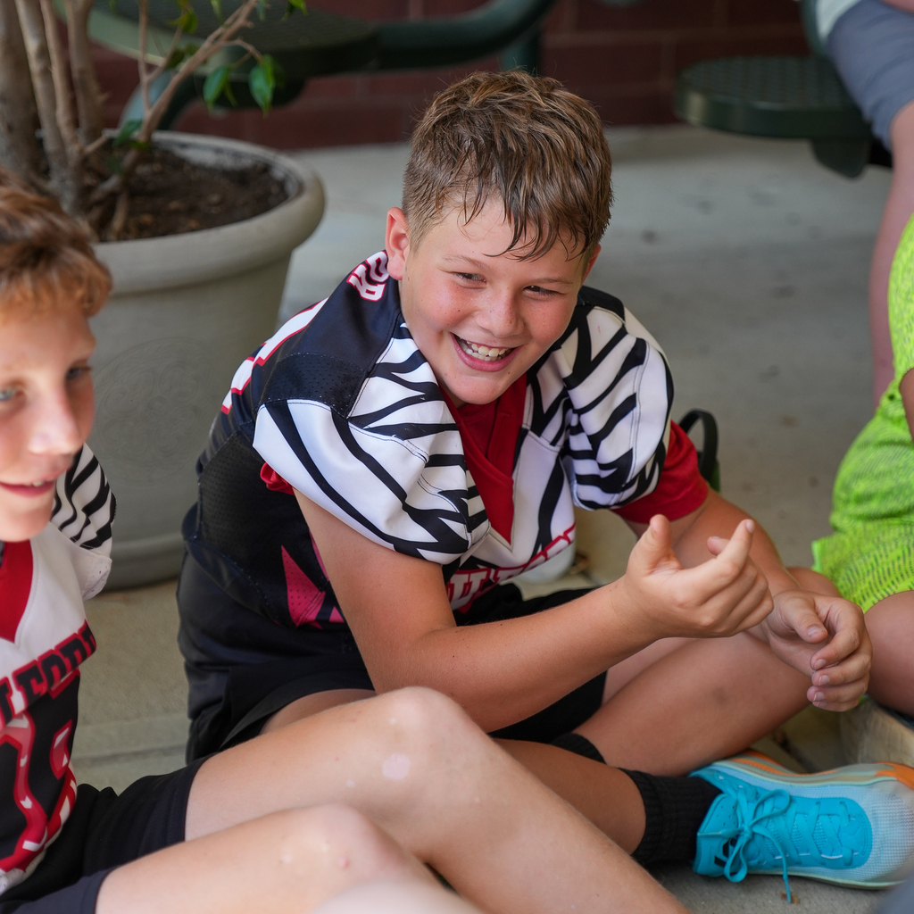 A McCormick Elementary student wearing a Milford jersey smiles while resting with classmates after an outdoor activity.