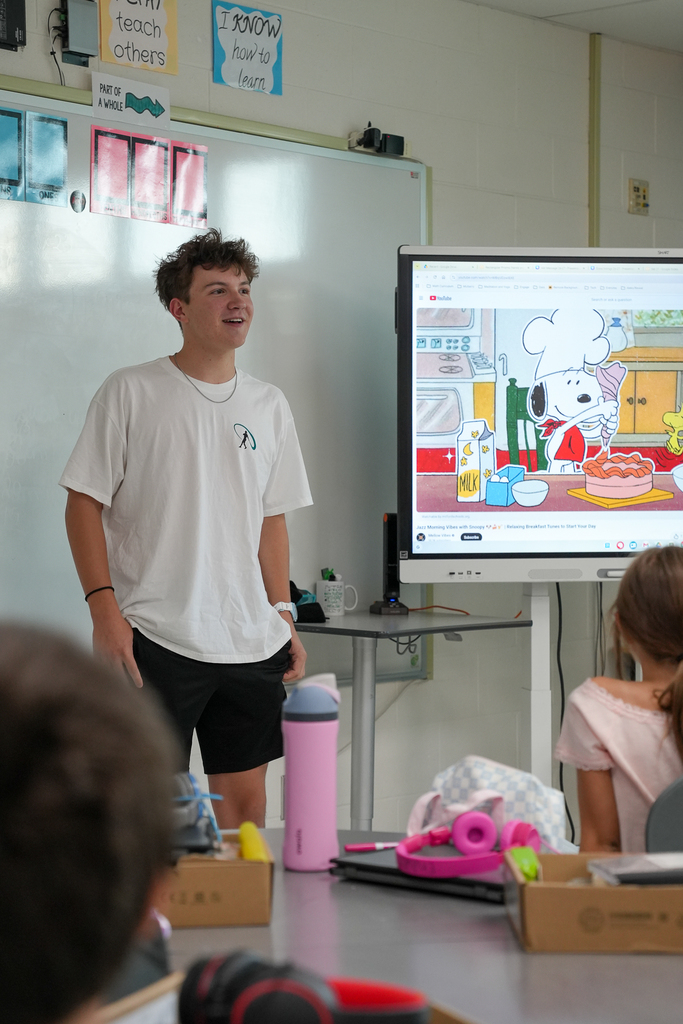 A Milford High School student mentor stands at the front of a Mulberry Elementary classroom speaking to students beside an interactive display.