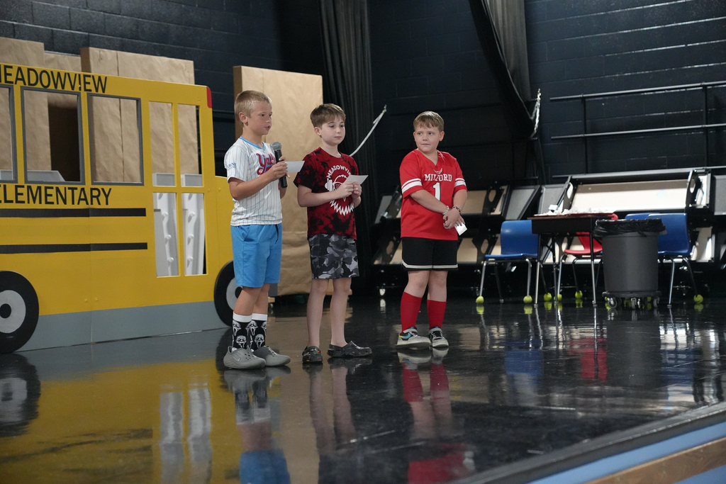 Three fifth-grade students speak onstage beside a large cardboard Meadowview Elementary school bus.