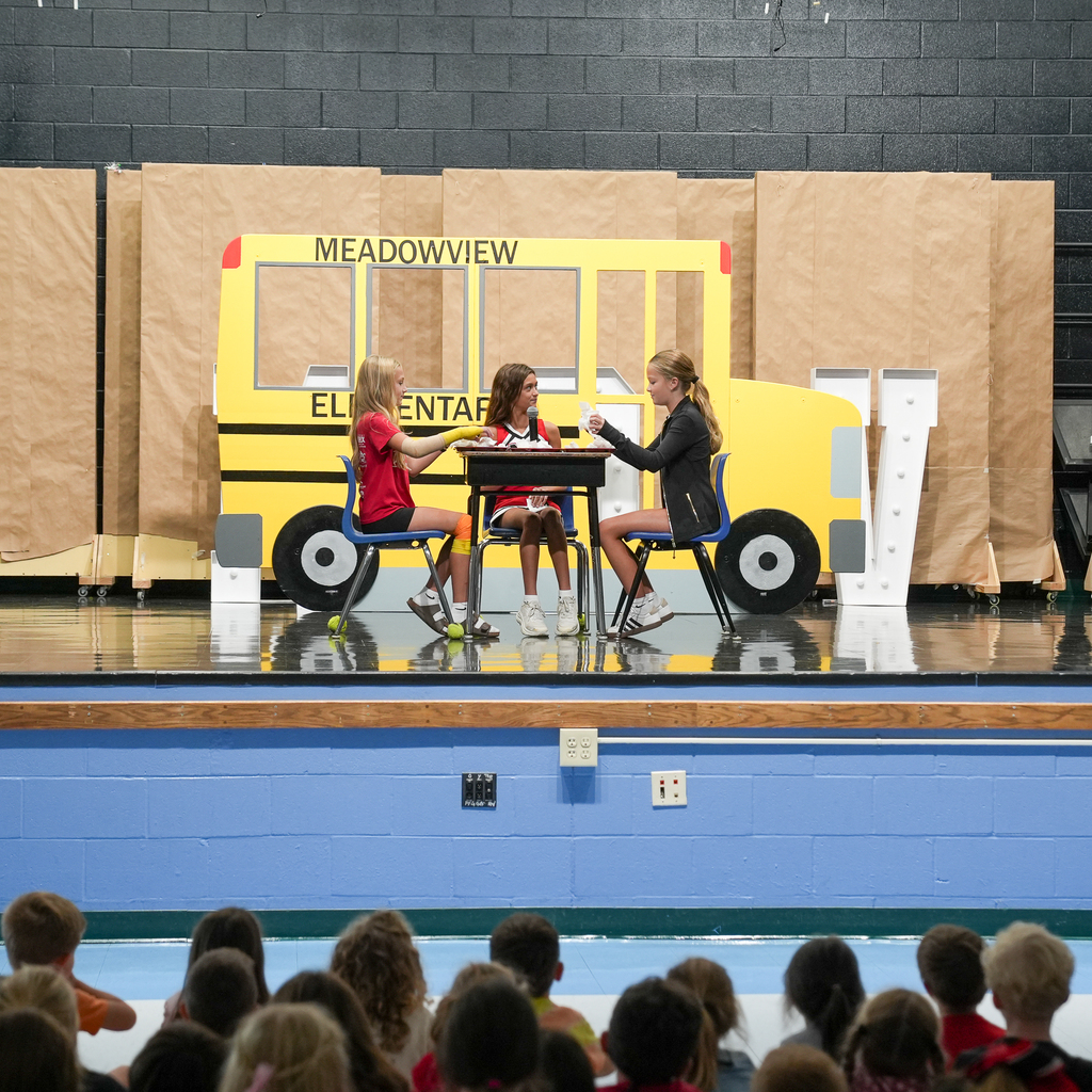 Three fifth-grade students act out a scenario onstage as two staff members watch nearby.