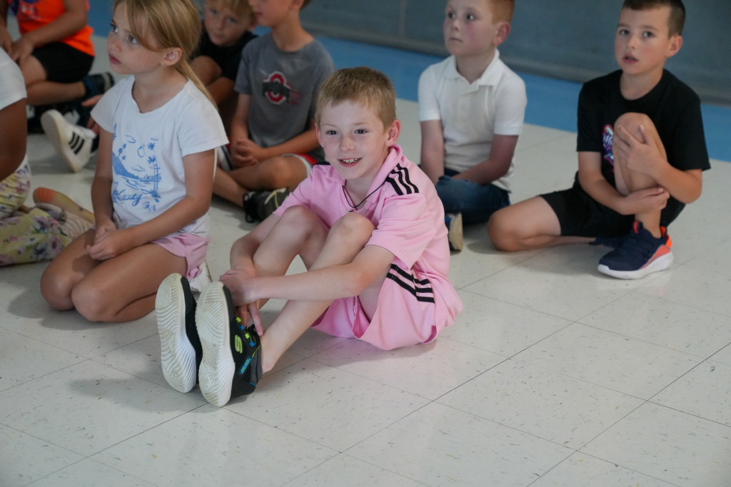 A smiling student in pink watches the assembly while seated with classmates on the gym floor.