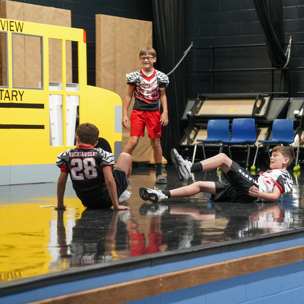 Three students wearing Milford football jerseys act out a behavior scenario onstage.
