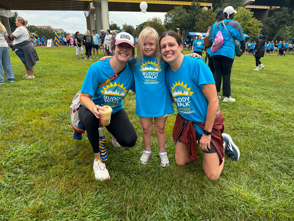 A child wearing a blue Cincinnati Buddy Walk shirt stands between two adults as they smile together on the grass at the event.