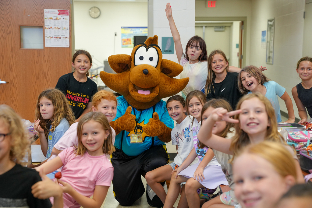 Munch the Fox gives two thumbs up while surrounded by a group of smiling elementary students in the cafeteria.