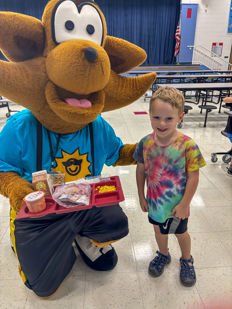 Munch the Fox kneels beside a smiling elementary student while holding a tray of school lunch items.