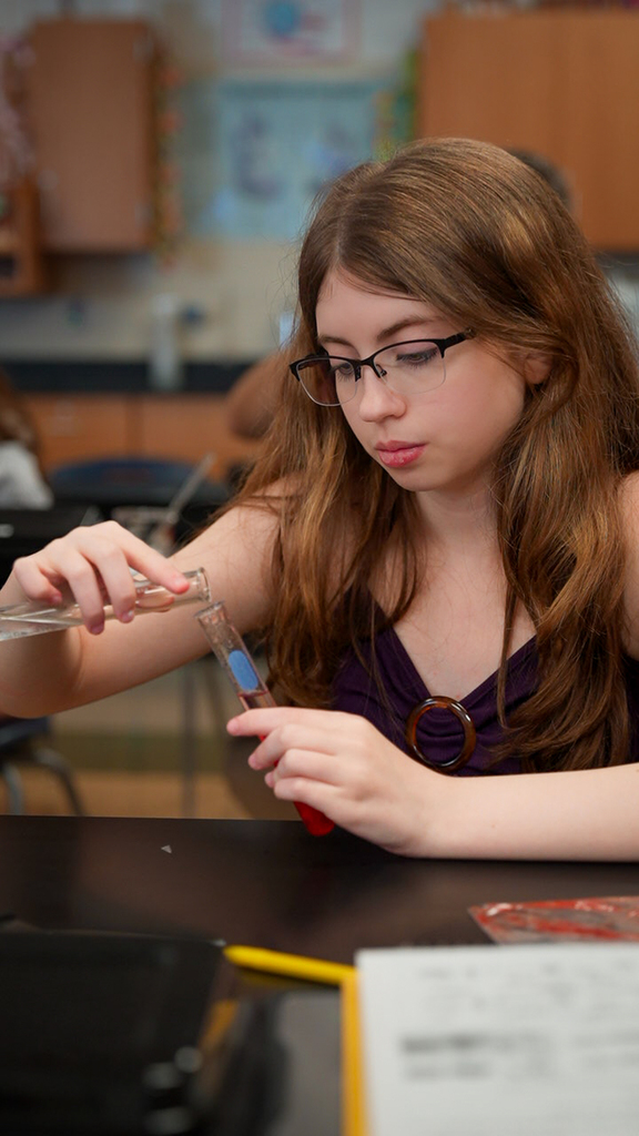 Students examine test tubes containing red strawberry mixtures while working at classroom lab tables during a DNA extraction activity.
