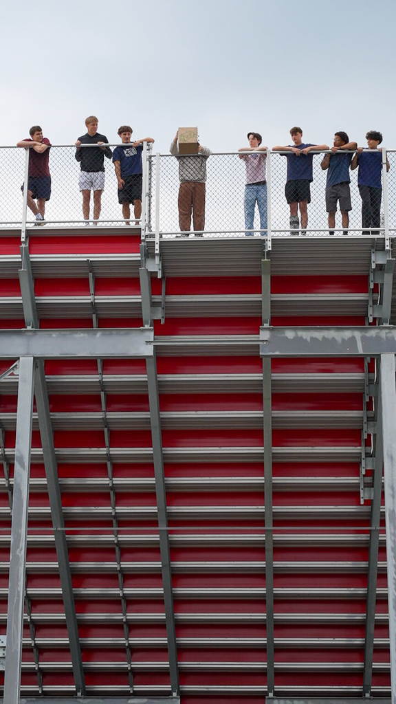 Students gather behind the railing at the top of the stadium bleachers as one holds a cardboard protective chamber in preparation for the egg drop.
