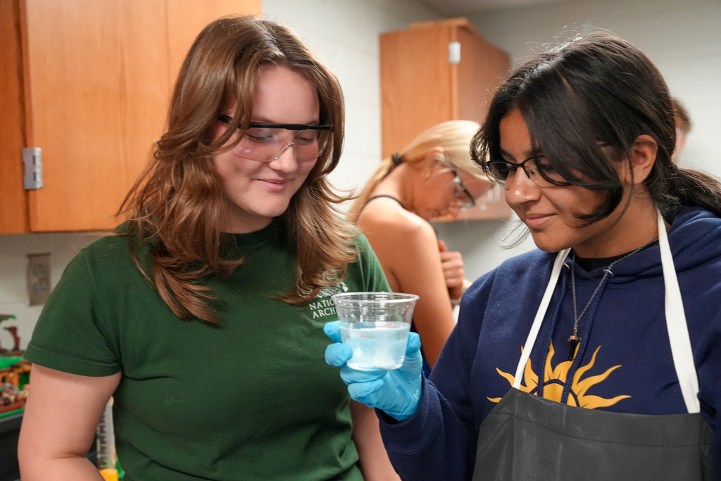 Two students observe a clear cup containing liquid and ice. One wears blue gloves and a lab apron and holds the cup up for a closer look.